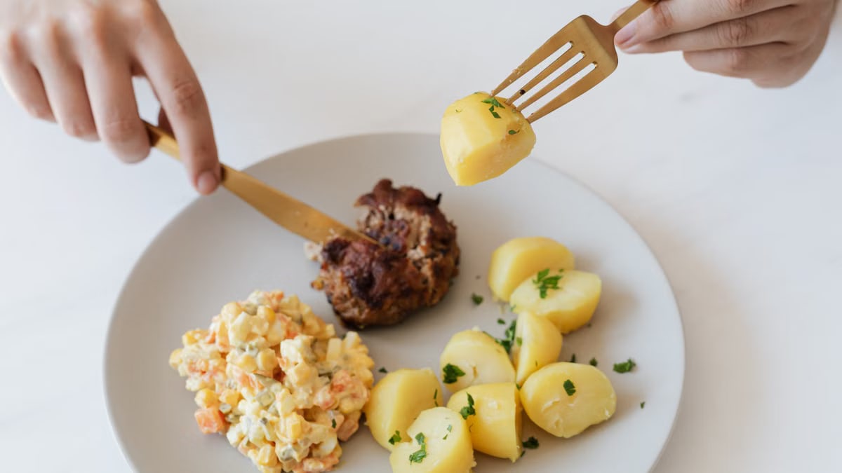 From above of crop anonymous person with cutleries eating delicious fried cutlet and hot boiled potatoes with parsley garnished with nutritious Russian salad